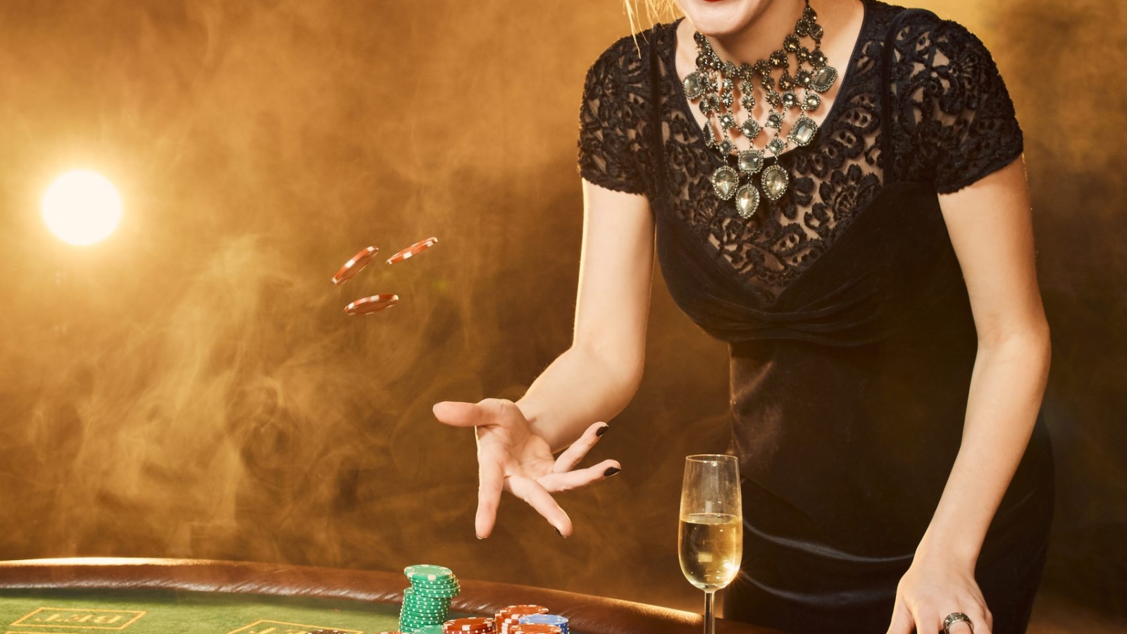 Young woman in evening dress with chips in hand standing near poker table with glass of champagne