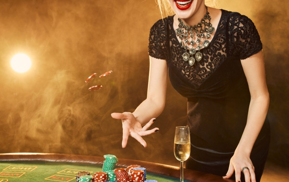 Young woman in evening dress with chips in hand standing near poker table with glass of champagne